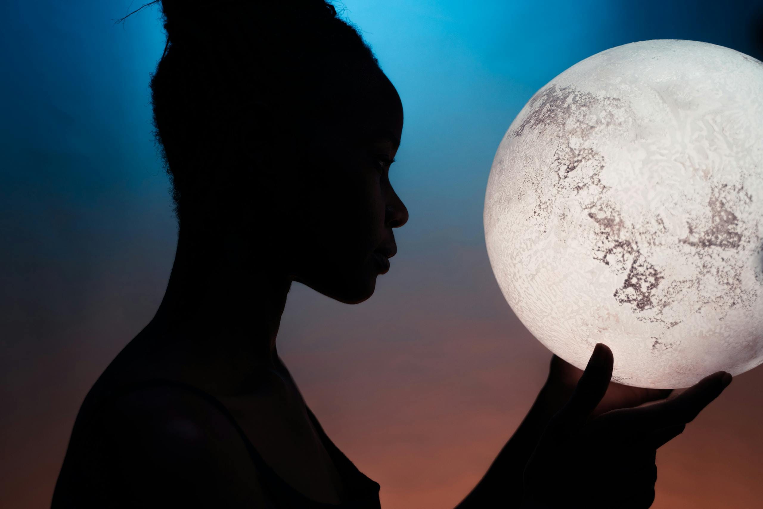 Artistic silhouette of woman holding a glowing moon globe against a vibrant backdrop.