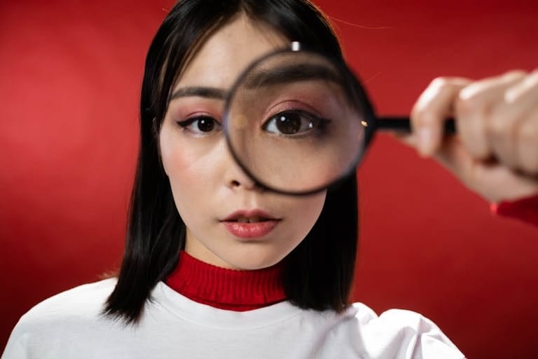 Asian woman holding magnifying glass over eye against red background.