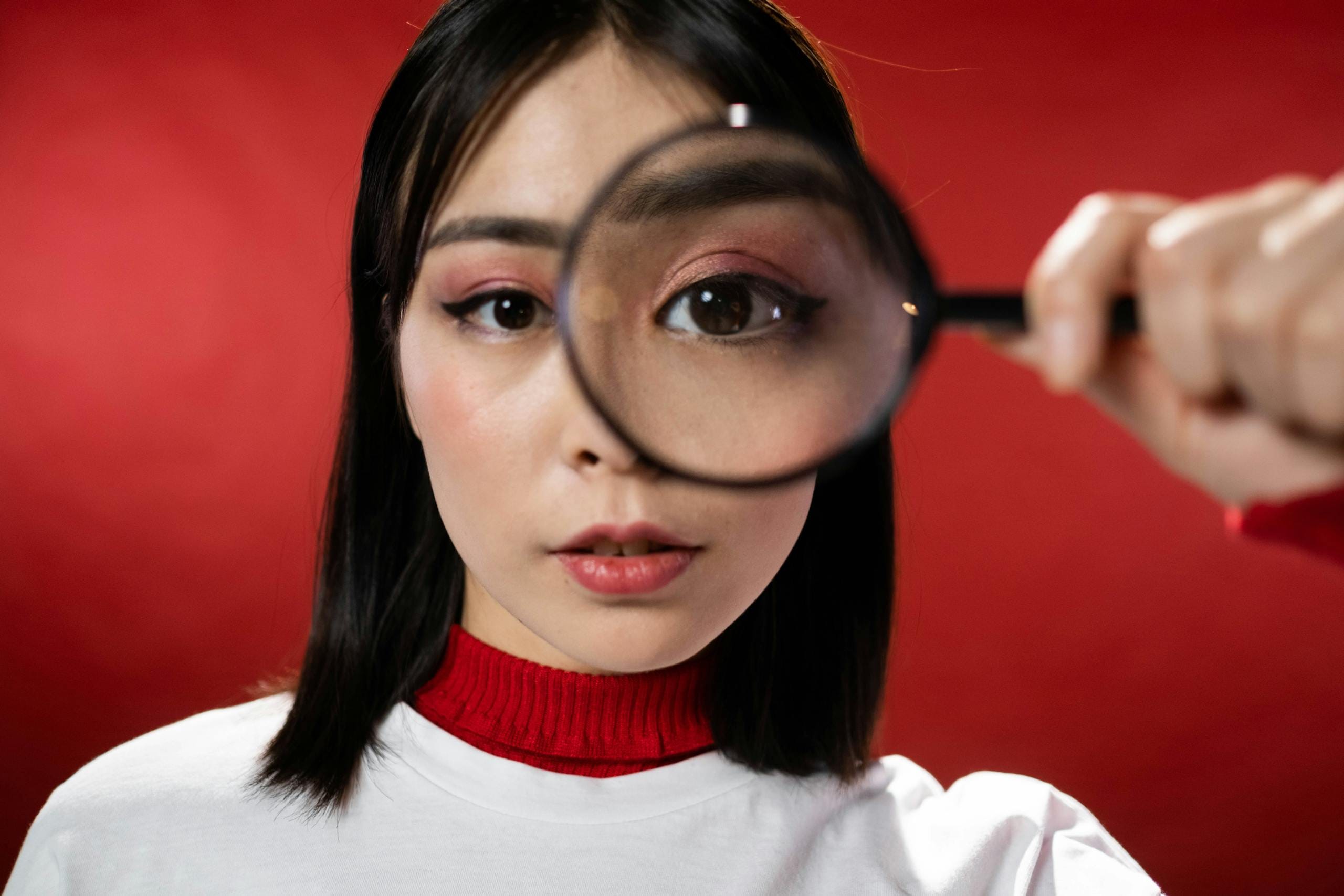 Asian woman holding magnifying glass over eye against red background.