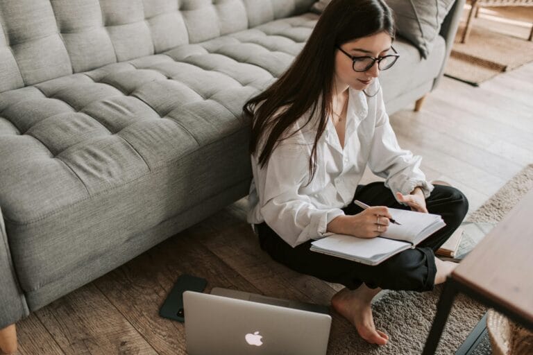 Entry-Level Remote Jobs That Pay Well A young woman sitting on the floor using a laptop and writing notes in a cozy apartment.