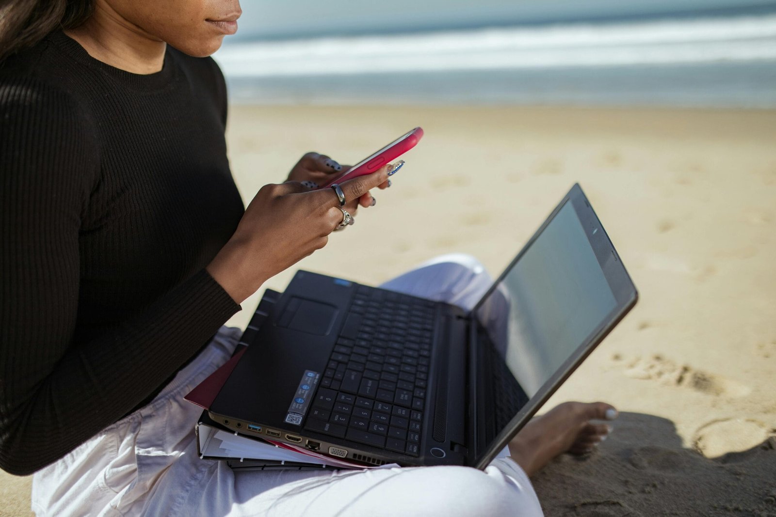 Person multitasking with a laptop and smartphone by the beach, embracing remote work lifestyle.
